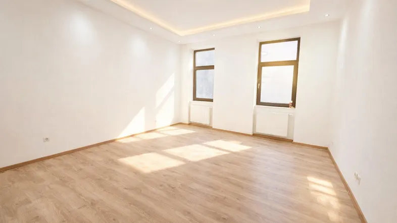 Helles, leeres Zimmer mit Laminatboden und zwei Fenstern, das viel Tageslicht hereinlässt.