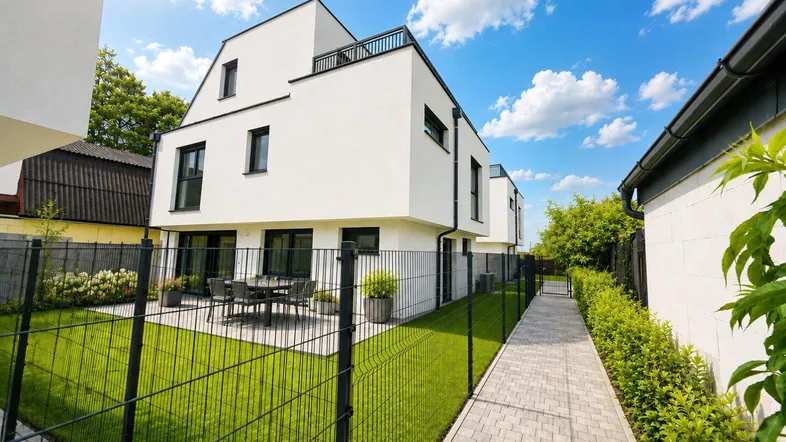 Modernes weißes Gebäude mit Terrasse und kleinem Gartenbereich unter strahlend blauem Himmel.