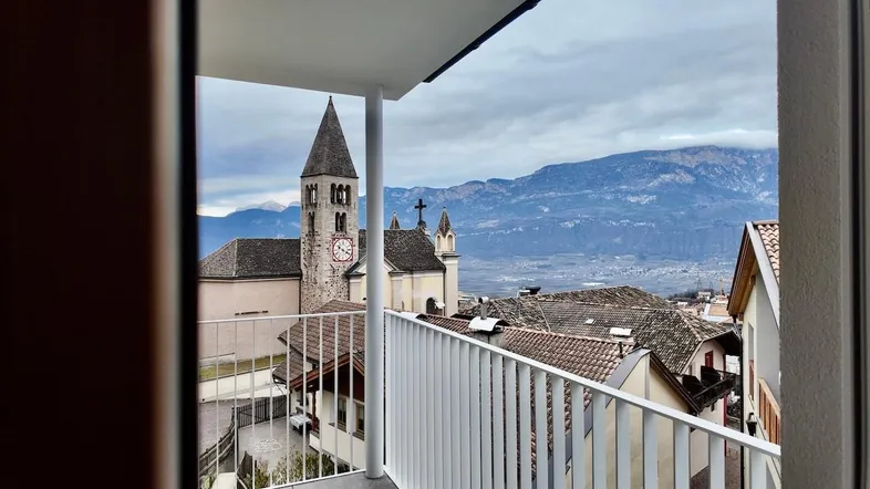 Blick vom Balkon auf eine Kirche und die umliegende Berglandschaft unter bewölktem Himmel.