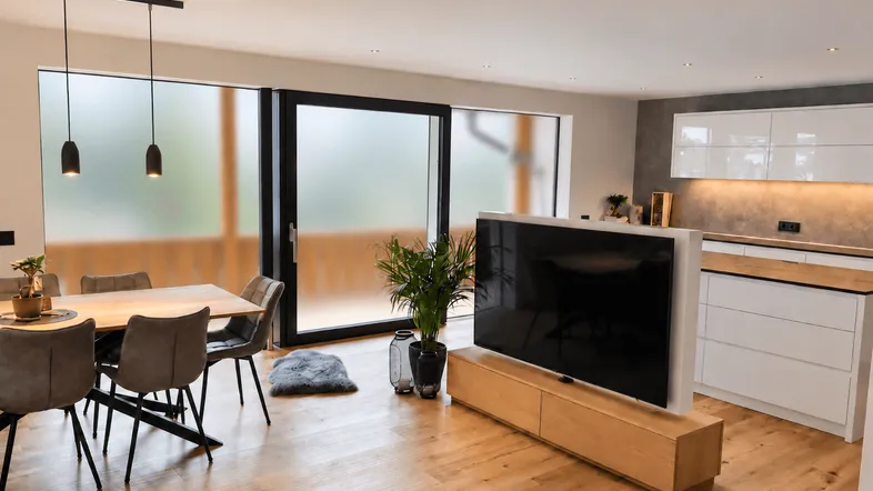 Moderner offener Wohn-Essbereich mit Holzboden, integrierter Küche und Zugang zu einem Balkon.