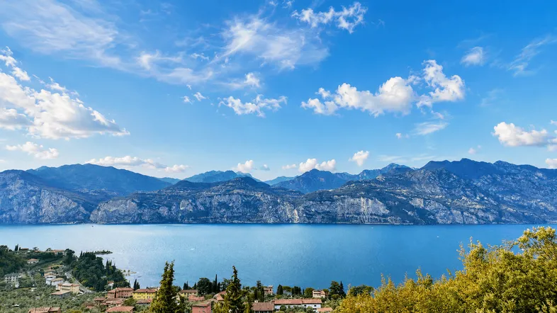 Panoramablick auf den Gardasee mit majestätischen Bergen und einer malerischen Uferlandschaft unter blauem Himmel.