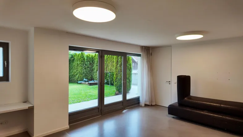 Heller Wohnbereich mit großen Glastüren, die zu einem grünen Garten führen, und einem dunklen Sofa.