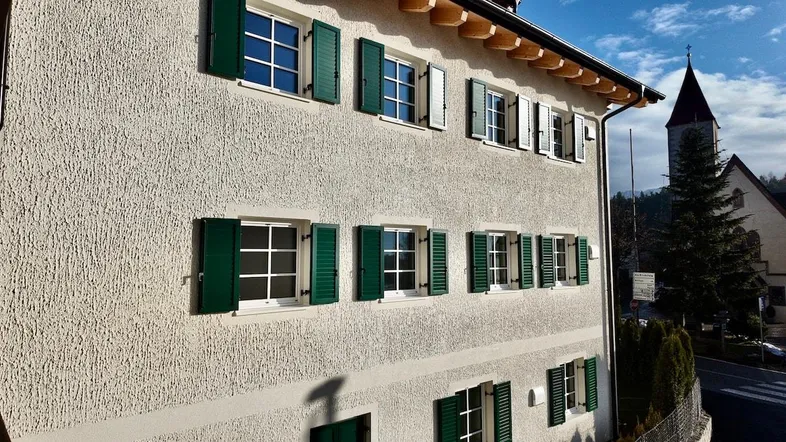 Traditionelles Gebäude mit grünen Fensterläden und einem Kirchturm im Hintergrund.