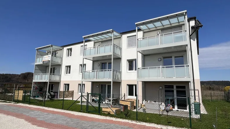 Modernes Mehrfamilienhaus mit mehreren Balkonen und klarem blauen Himmel, umgeben von Grünflächen.