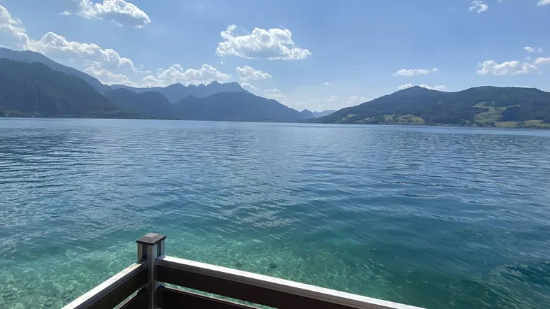 Panoramablick über den See und die umliegenden Berge von einem Balkon aus, unter blauem Himmel.