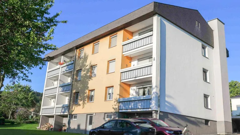 Mehrfamilienhaus mit farbiger Fassade, mehreren Balkonen und umliegender Grünfläche unter blauem Himmel.