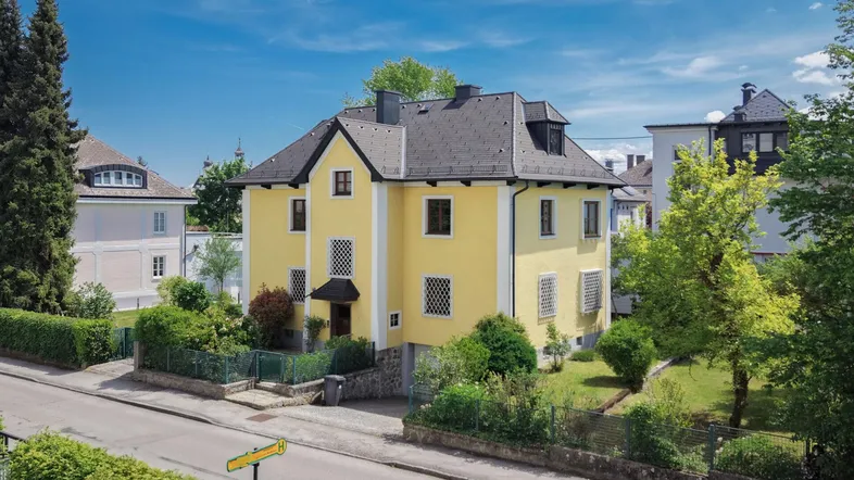Gelbe Villa mit Satteldach und gepflegtem Vorgarten unter blauem Himmel.