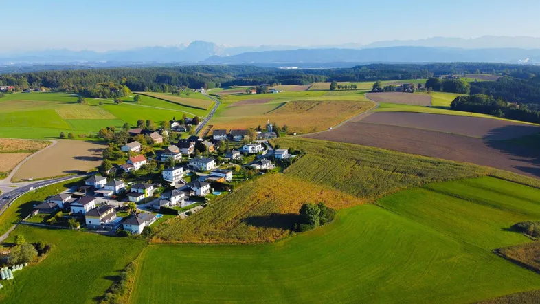 Luftaufnahme eines ländlichen Dorfes umgeben von grünen Feldern mit Bergen im Hintergrund.