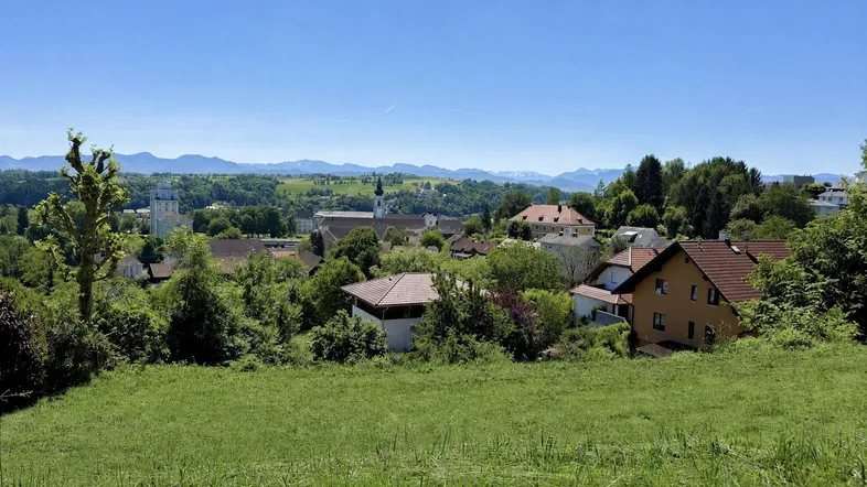 Panoramablick über ein grünes Dorf mit Häusern, Bäumen und einer Kirche im Hintergrund, umgeben von Bergen.