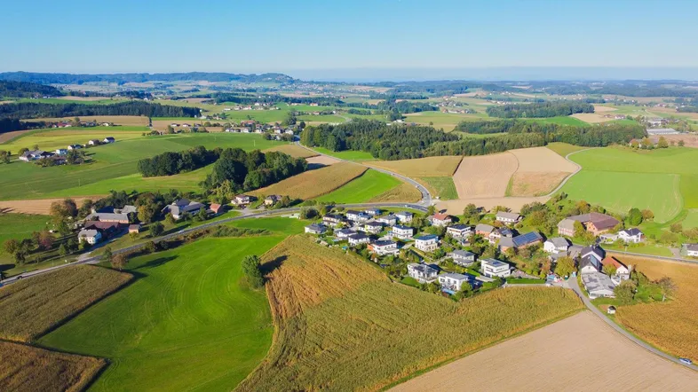 Luftaufnahme einer ländlichen Wohnsiedlung umgeben von Feldern und grünen Hügeln.