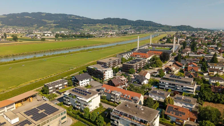 Luftaufnahme einer Wohnsiedlung mit grünen Feldern, einem Fluss und Bergen im Hintergrund.