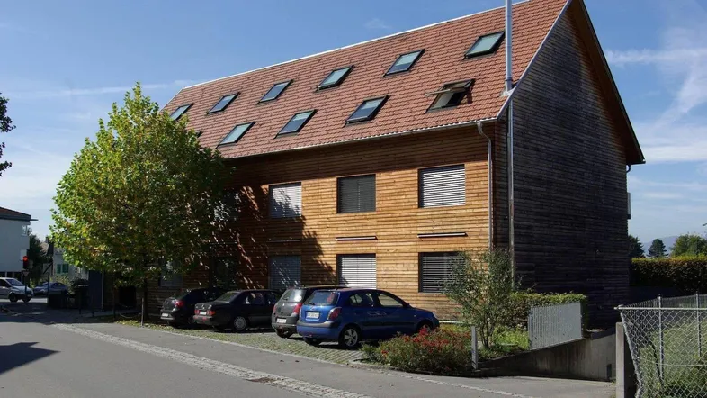 Modernes Gebäude mit Holzfassade, rotem Ziegeldach und vielen Dachfenstern, Parkplätze davor.