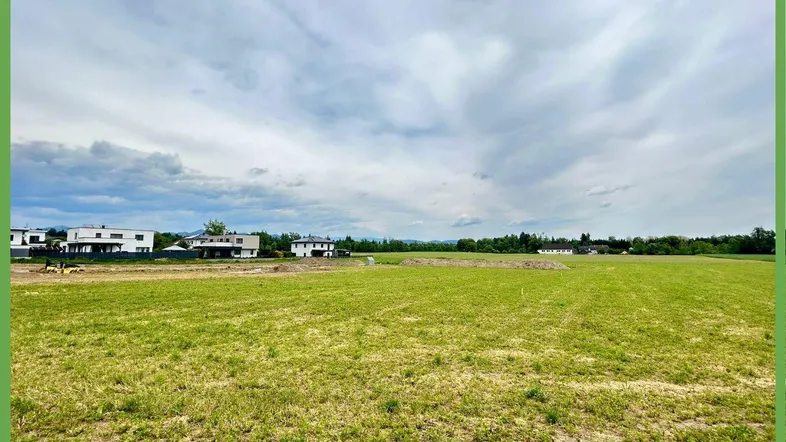 Weitläufiges grünes Feld mit modernen Häusern in der Ferne unter bewölktem Himmel.