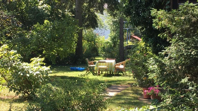 Idyllischer Gartenbereich mit Sitzgelegenheiten unter schattenspendenden Bäumen und blühenden Pflanzen.