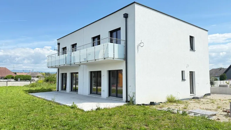 Modernes weißes Gebäude mit Balkonen und einer Terrasse auf einem unbebauten Grundstück.