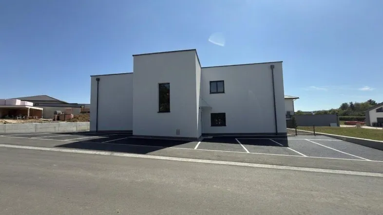 Moderne, weiß verputzte Fassade eines Neubaus mit Flachdach und zugehörigen Parkplätzen unter blauem Himmel.