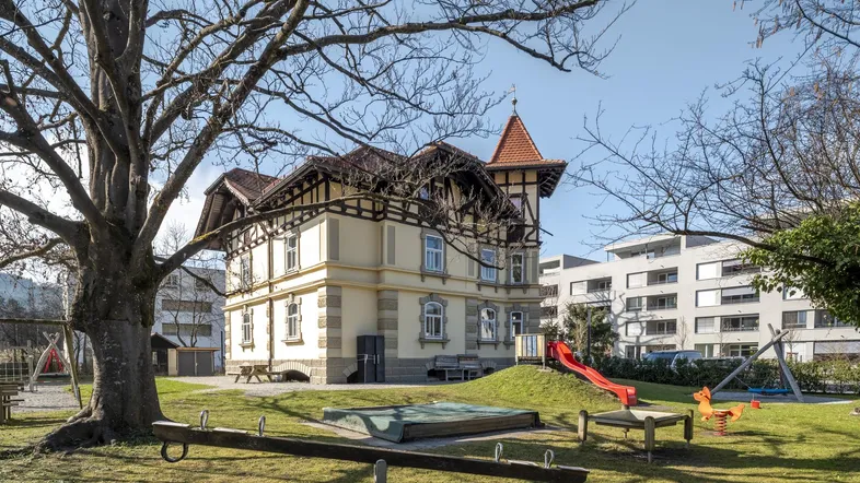 Historisches Gebäude mit Fachwerkfassade und einem Spielplatz im Vordergrund unter blauem Himmel.