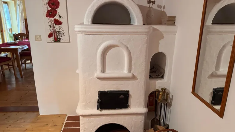 Traditioneller weißer Kachelofen in einem gemütlichen Wohnbereich mit Holzboden und Blick auf den Essbereich.