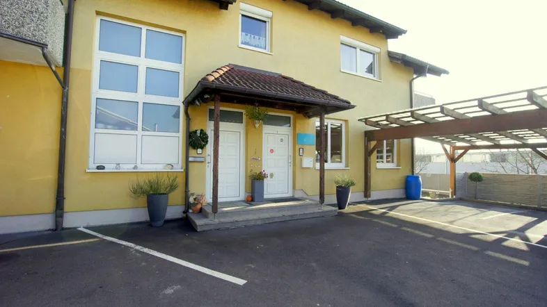 Gesamtansicht des gelben Gebäudes mit Carport und Eingangsbereich unter hellem Tageslicht.