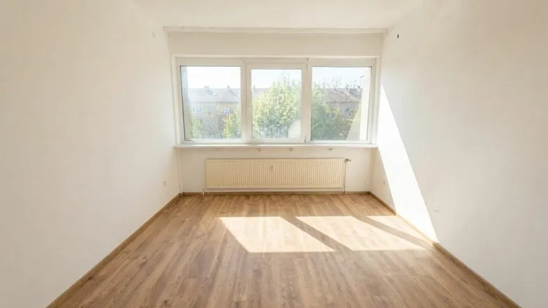 Heller Raum mit Laminatboden, weißen Wänden und Blick ins Grüne durch große Fenster.