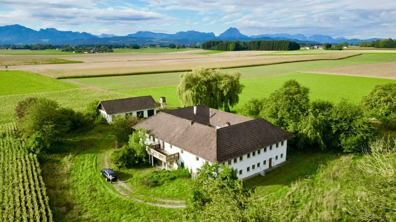 Luftaufnahme eines großen Landhauses umgeben von grünen Feldern mit Bergpanorama im Hintergrund.