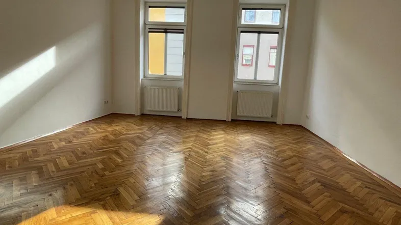 Helles Zimmer mit Fischgrätparkettboden und zwei Fenstern, die viel Tageslicht hereinlassen.