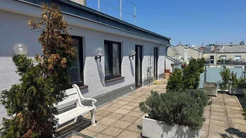 Große Dachterrasse mit Fliesenboden, einer weißen Sitzbank und Pflanzen in Kübeln unter blauem Himmel.
