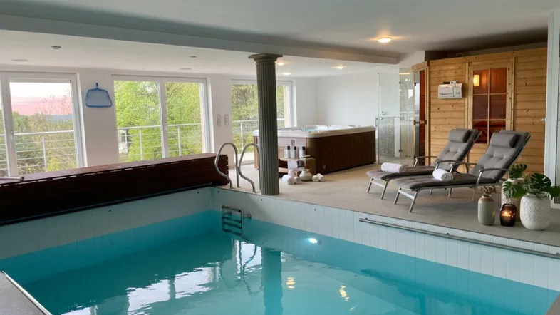 Großzügiger Wellnessbereich mit Innenpool, Whirlpool und Sauna, umgeben von großen Fenstern mit grüner Aussicht.