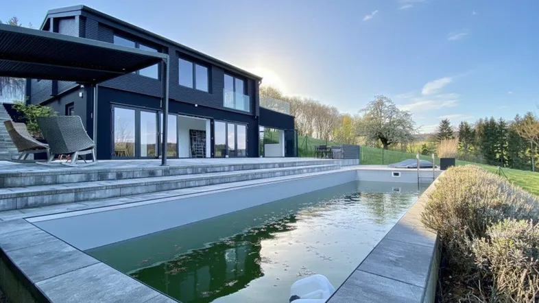 Modernes Haus mit schwarzer Fassade, großem Pool und weitläufiger Terrasse in sonniger Landschaft.