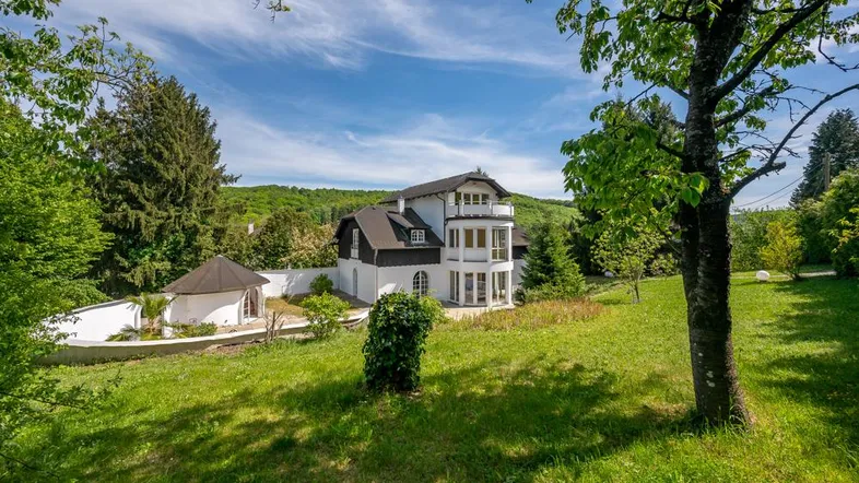 Malerische Villa mit weißer Fassade und dunklem Dach, eingebettet in eine grüne Landschaft unter blauem Himmel.