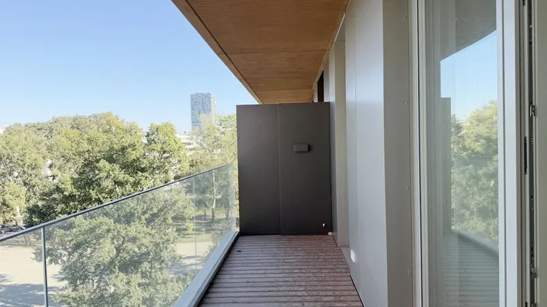 Geräumiger Balkon mit Holzboden und Glasgeländer bietet einen weiten Blick ins Grüne und auf die Stadt.