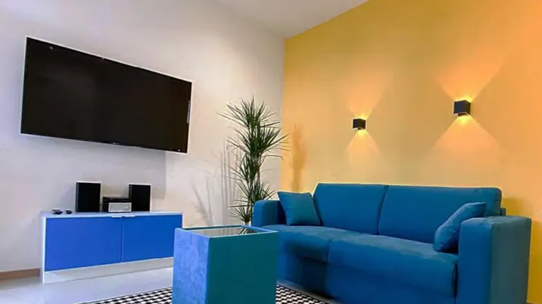 Helles Wohnzimmer mit einem blauen Sofa, gelber Akzentwand und modernem Flachbildfernseher.