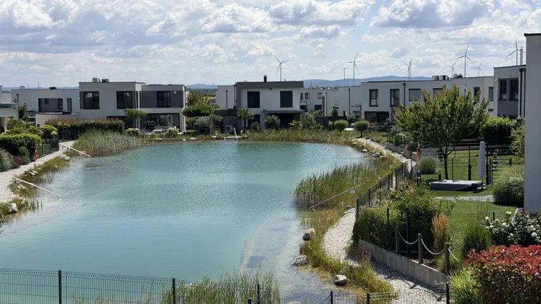 Blick auf die moderne Wohnanlage mit einem natürlichen Teich und Windrädern im Hintergrund.