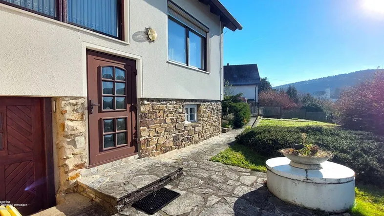 Eingangsbereich des Hauses mit Steinsockel und gepflastertem Weg an einem sonnigen Tag.
