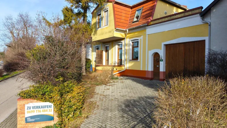 Gelbes Haus mit rotem Ziegeldach, Einfahrt und einem 'Zu Verkaufen'-Schild im Vorgarten.