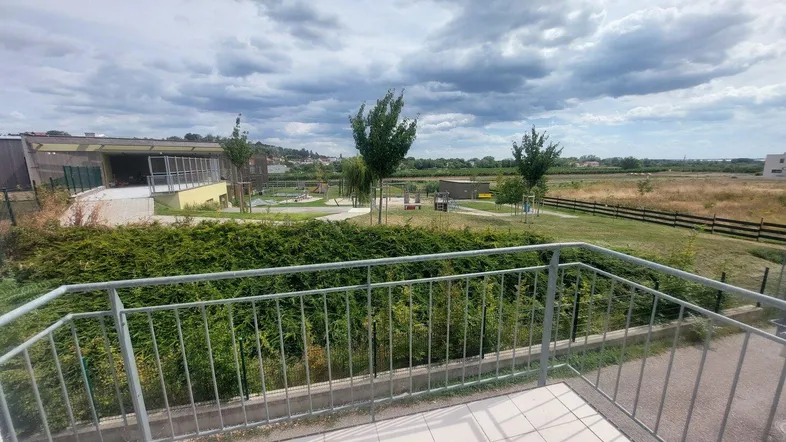 Blick von einem Balkon über eine Hecke auf eine weite Landschaft mit Spielplatz und Feldern.