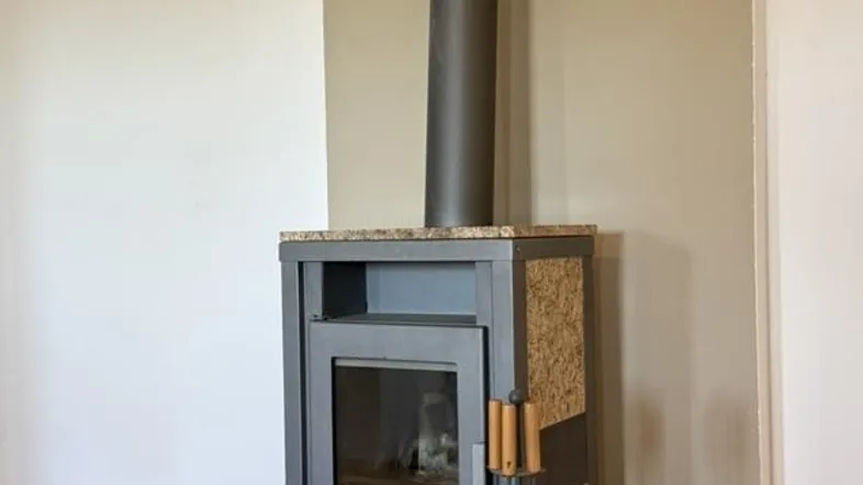 Moderner Holzofen mit Rauchrohr und Holzkorb auf Parkettboden in einem hellen Wohnbereich.