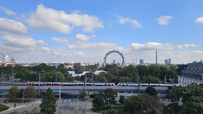 Weitläufiger Stadtblick mit dem Wiener Riesenrad und vorbeifahrendem Zug unter blauem Himmel.