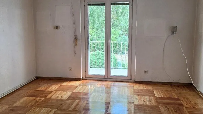 Helles Zimmer mit glänzendem Parkettboden und Zugang zu einem Balkon mit Grünblick.