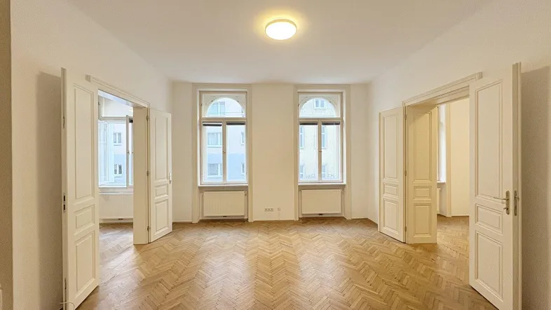 Geräumiges Zimmer mit Fischgrätparkett und hohen Fenstern, das viel Tageslicht hereinlässt.