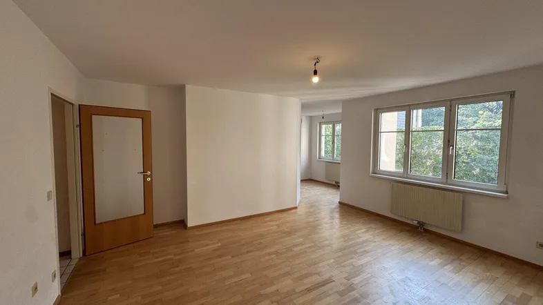 Helles, leeres Wohnzimmer mit Parkettboden und großen Fenstern, die viel Tageslicht hereinlassen.