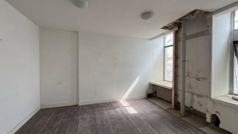 Unfertiger Innenraum mit dunklem Holzfußboden, weißen Wänden und sichtbaren Renovierungsspuren an den Fenstern.