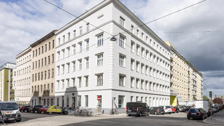 Ansicht eines Eckgebäudes in der Stadt mit mehreren Stockwerken und vielen Fenstern, umgeben von parkenden Autos.