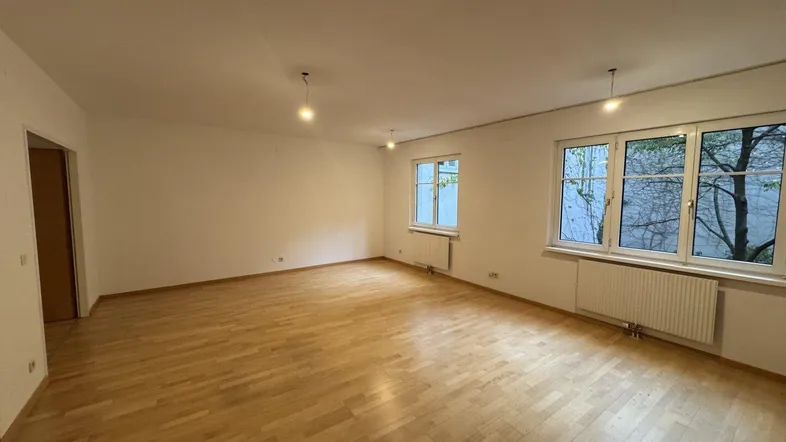 Geräumiges Wohnzimmer mit hellem Parkettboden und großen Fenstern, die viel Tageslicht hereinlassen.