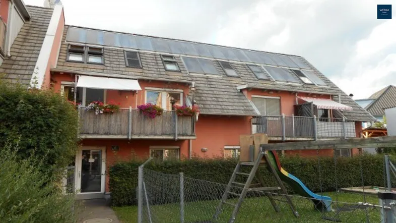 Mehrfamilienhaus mit Photovoltaikanlage auf dem Dach, Balkonen und einem Spielplatz im Gartenbereich.