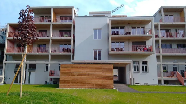 Modernes Mehrfamilienhaus mit Balkonen und einer Holzverkleidung im Erdgeschoss, umgeben von einer Grünfläche.
