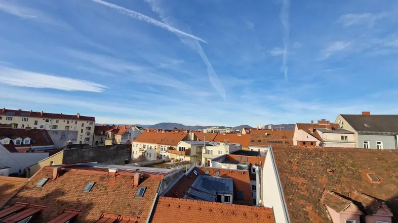 Weitläufiger Blick über die Dächer einer Stadt mit roten Ziegeln und Bergen im Hintergrund unter blauem Himmel.