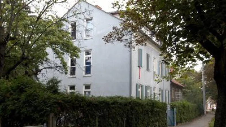 Gebäudeansicht eines gepflegten Altbaus mit hellgrauer Fassade und umgebender Hecke.