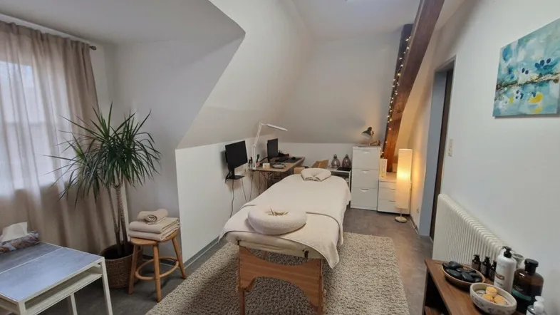 Heller Dachbodenraum, eingerichtet als Behandlungszimmer, mit schrägen Wänden, Dachfenster und sichtbarem Holzbalken.