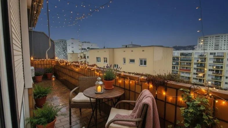 Gemütlicher Balkon am Abend mit Lichterketten, Sitzgelegenheiten und Blick auf die Stadt.